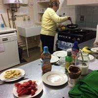 Doris in the background tending to a pancaake in a frying pan, while a selection of toppings and sauces is on a  counter top in the foreground.
